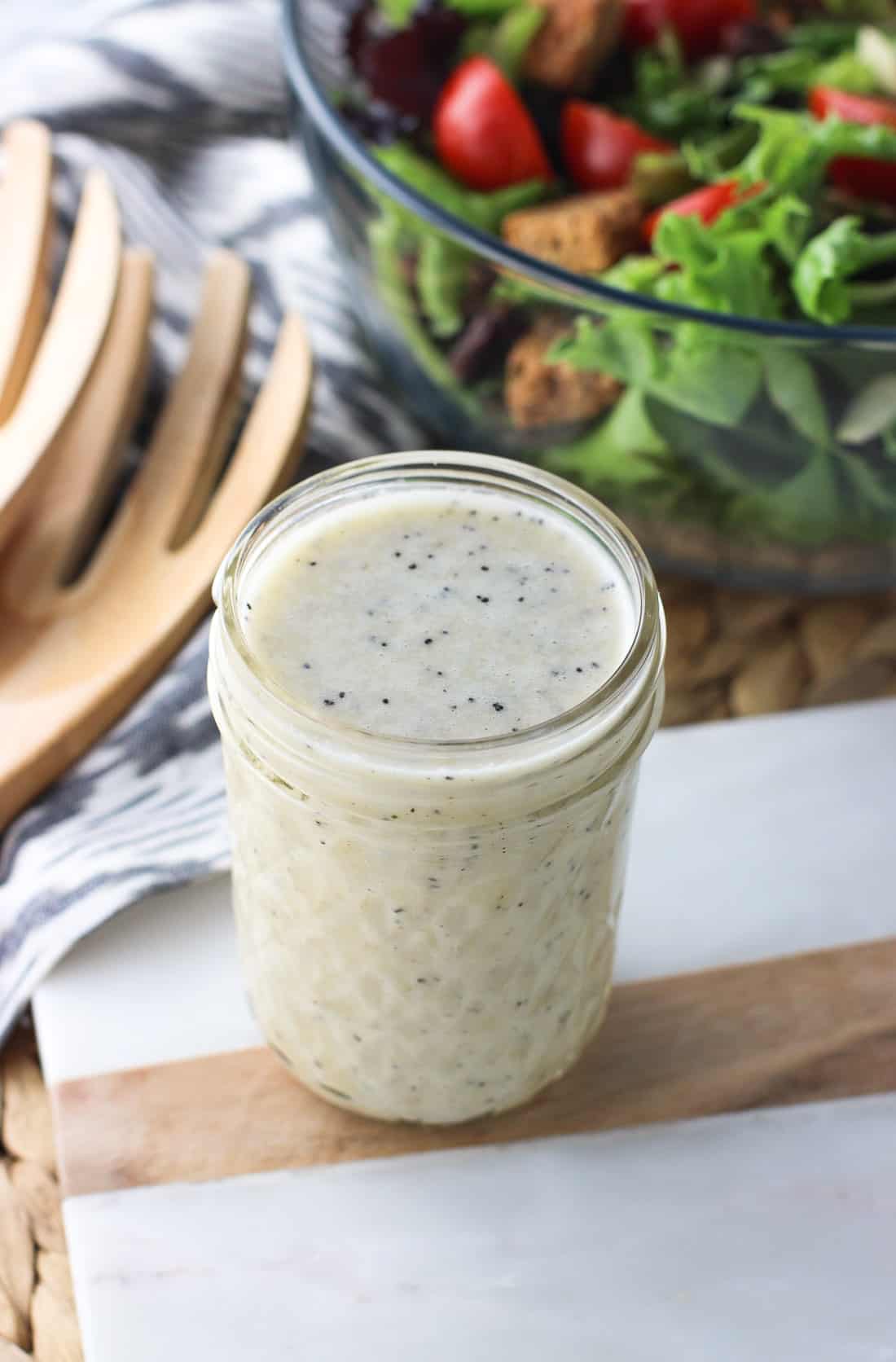 A jar of sweet onion dressing on a marble serving board in front of a big bowl of salad and wooden tongs