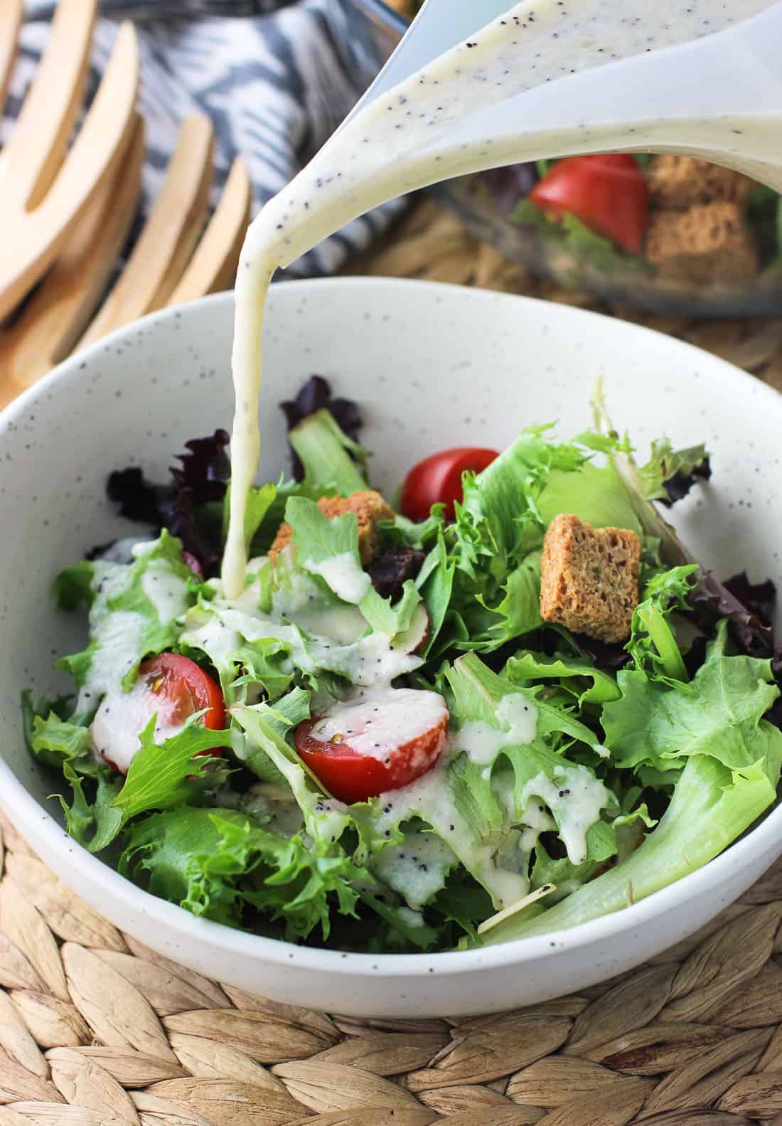 Dressing being poured over a shallow bowl of salad consisting of mixed greens, cherry tomatoes, and croutons