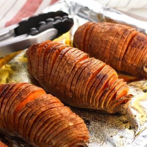 Hasselback cut sweet potatoes on an aluminum foil-lined baking sheet with tongs in the background