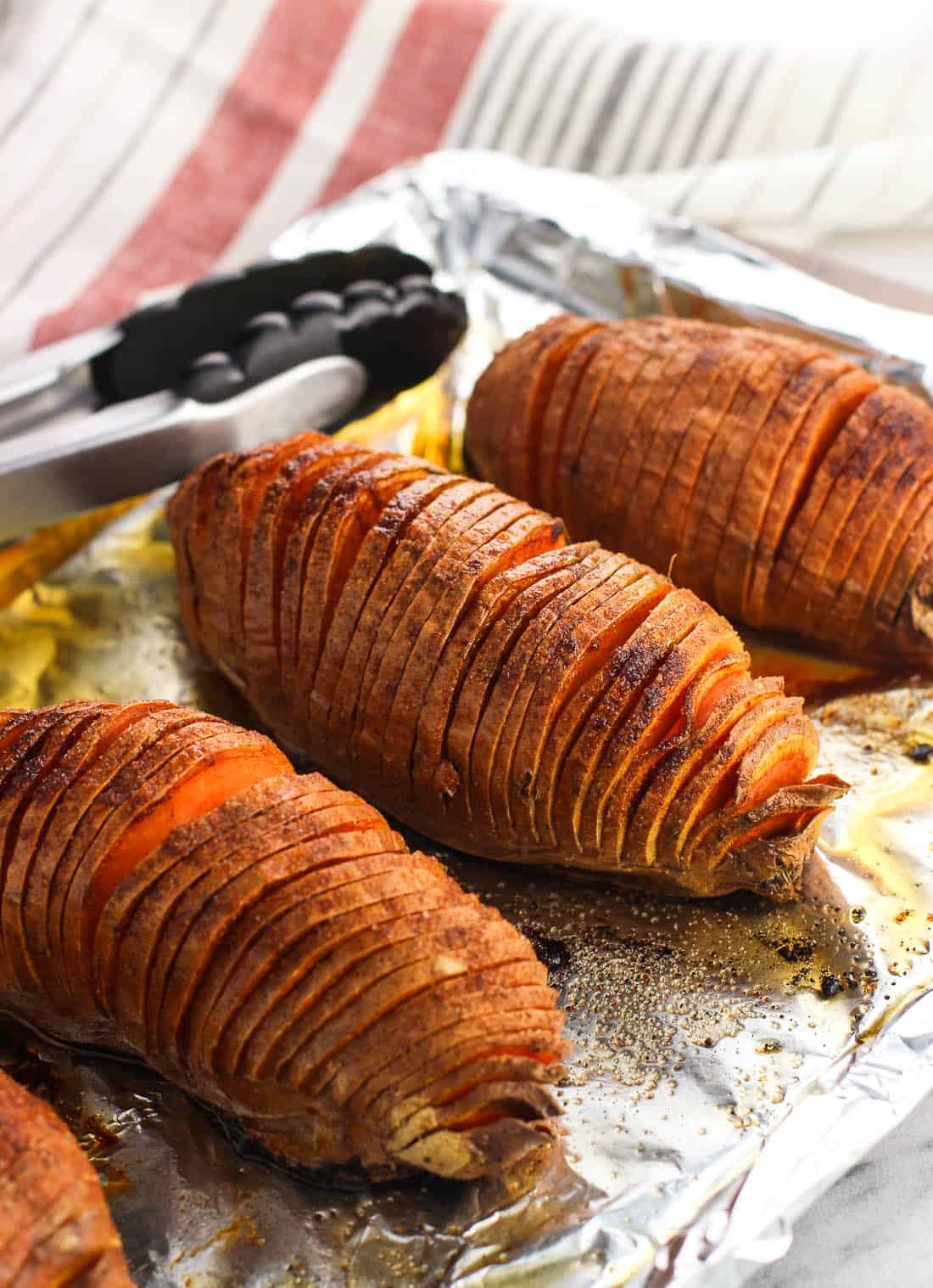 Hasselback cut sweet potatoes on an aluminum foil-lined baking sheet with tongs in the background