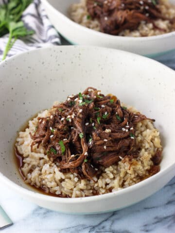A pile of shredded beef served over brown rice in a shallow dinner bowl