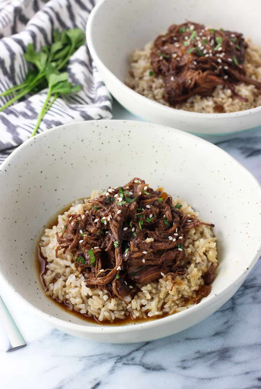 A bowl of shredded beef served over brown rice garnished with sesame seeds, herbs, and sauce with another bowl in the background