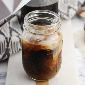 A ceramic milk carton hovering above a mason jar of coffee swirled with milk after pouring some in