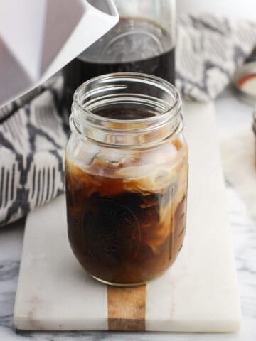 A ceramic milk carton hovering above a mason jar of coffee swirled with milk after pouring some in