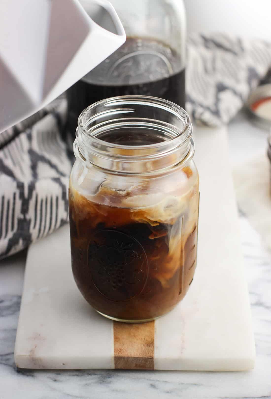 A ceramic milk carton hovering above a mason jar of coffee swirled with milk after pouring some in.