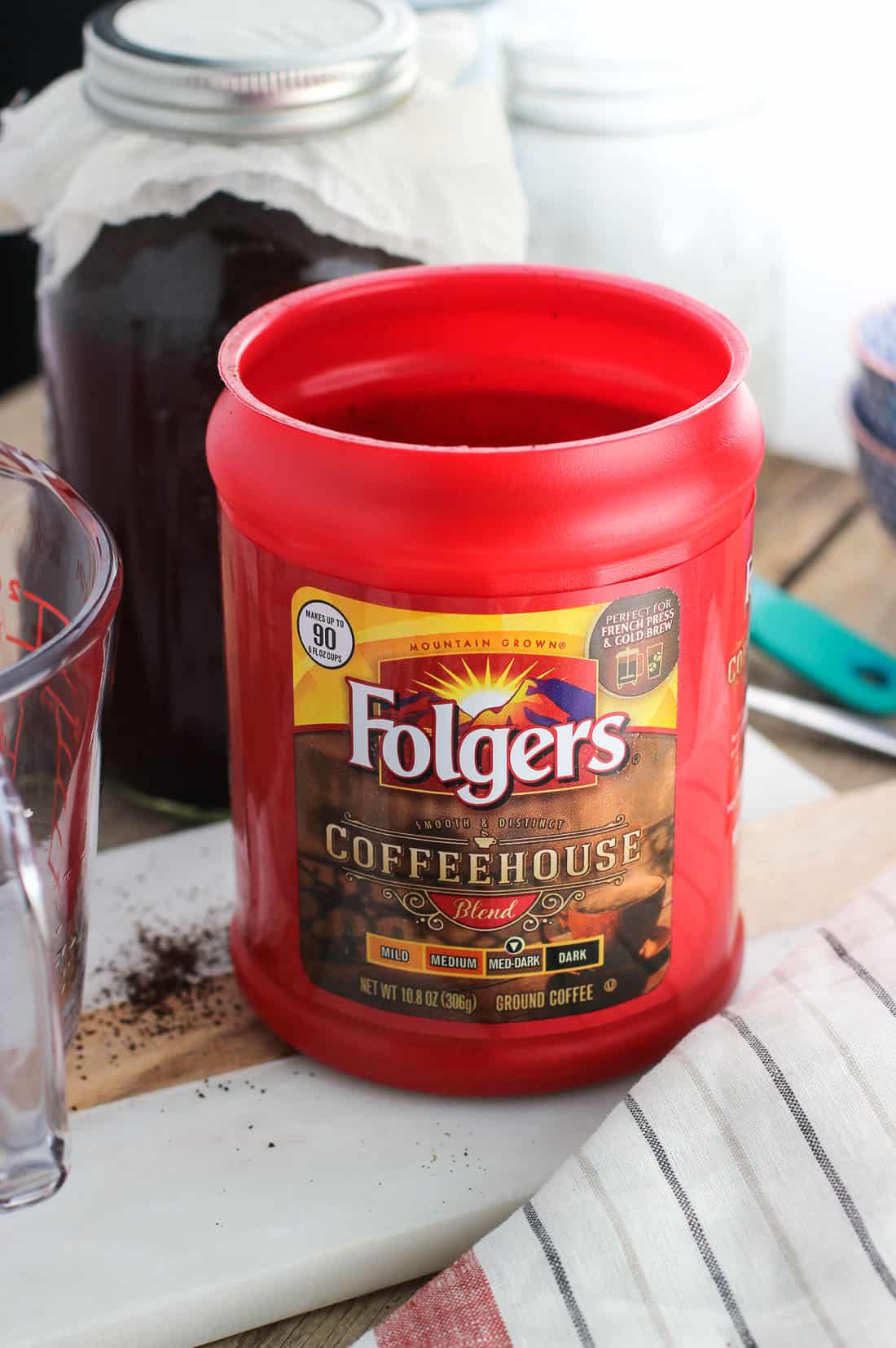 A plastic carton of Folgers Coffeehouse Blend ground coffee on a board in front of a jar of cold brew coffee.