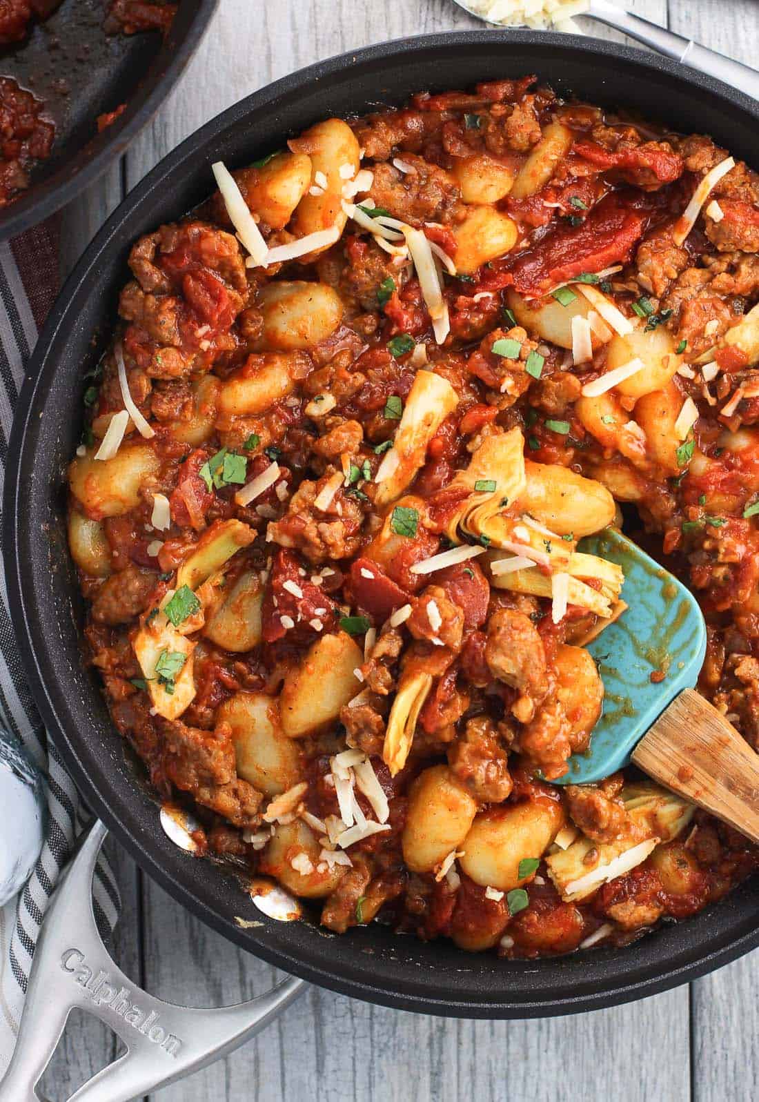 An overhead picture of a skillet full of gnocchi, crumbled Italian sausage, marinara, and artichoke hearts with a spatula in it