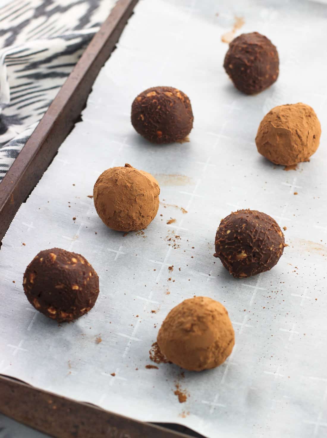 Seven truffles on a parchment-lined baking sheet, with three truffles having been rolled in cocoa powder