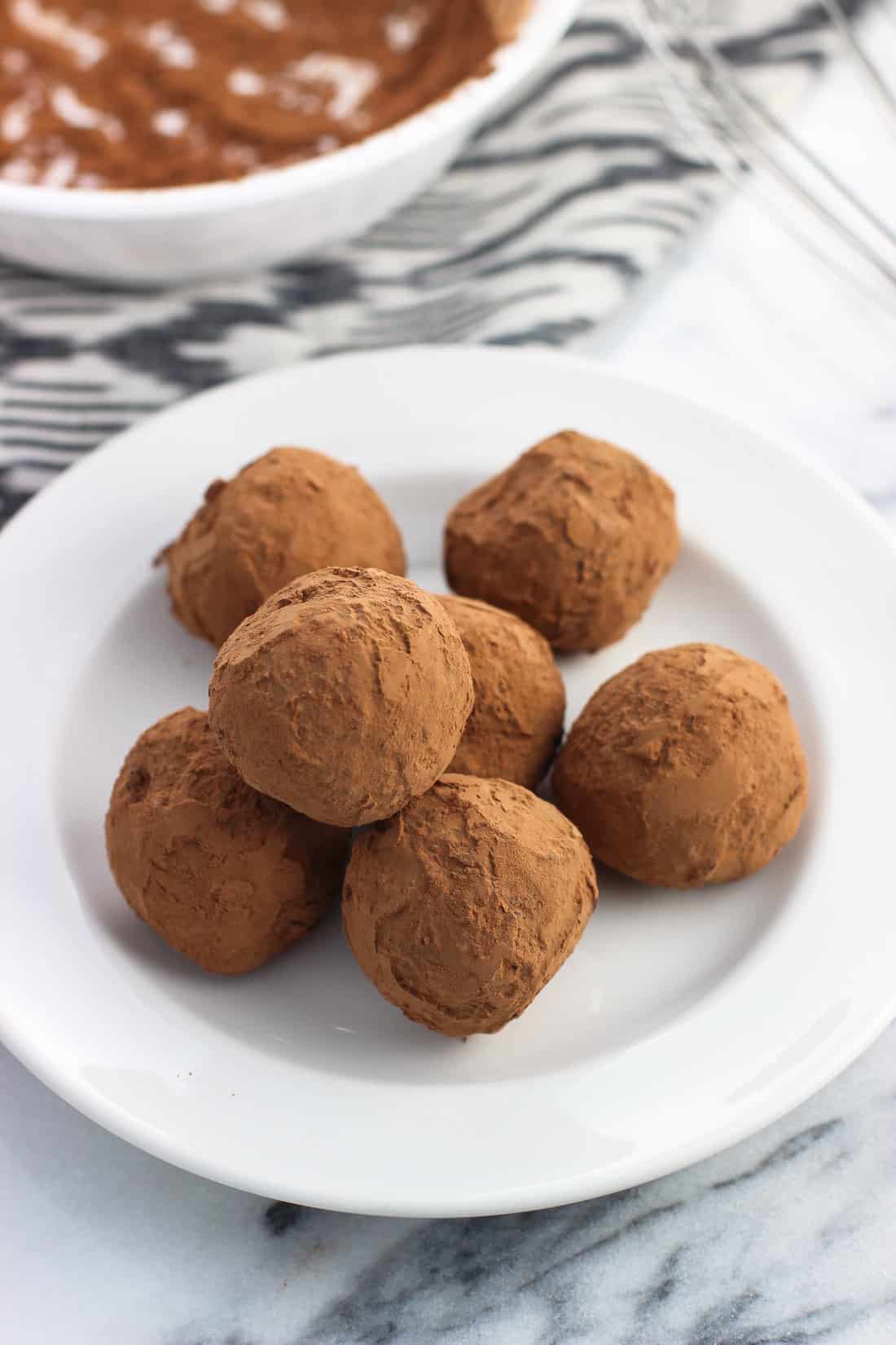 Seven truffles piled on a ceramic dessert plate with a bowl of cocoa powder and a whisk in the background