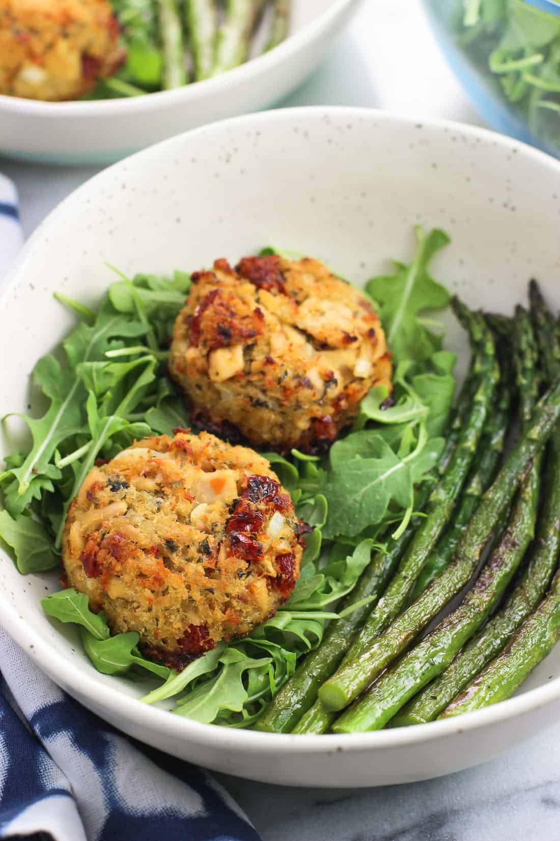 A close-up of two tuna cakes in a dish with arugula and sauteed asparagus spears.