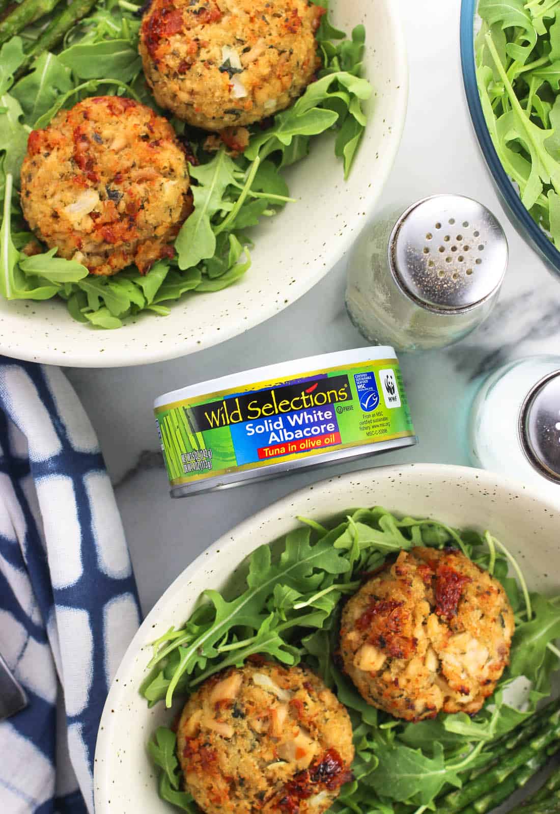 An overhead shot of two plates of two tuna cakes each served over a bed of arugula. A Wild Selections can of tuna is in focus between the plates