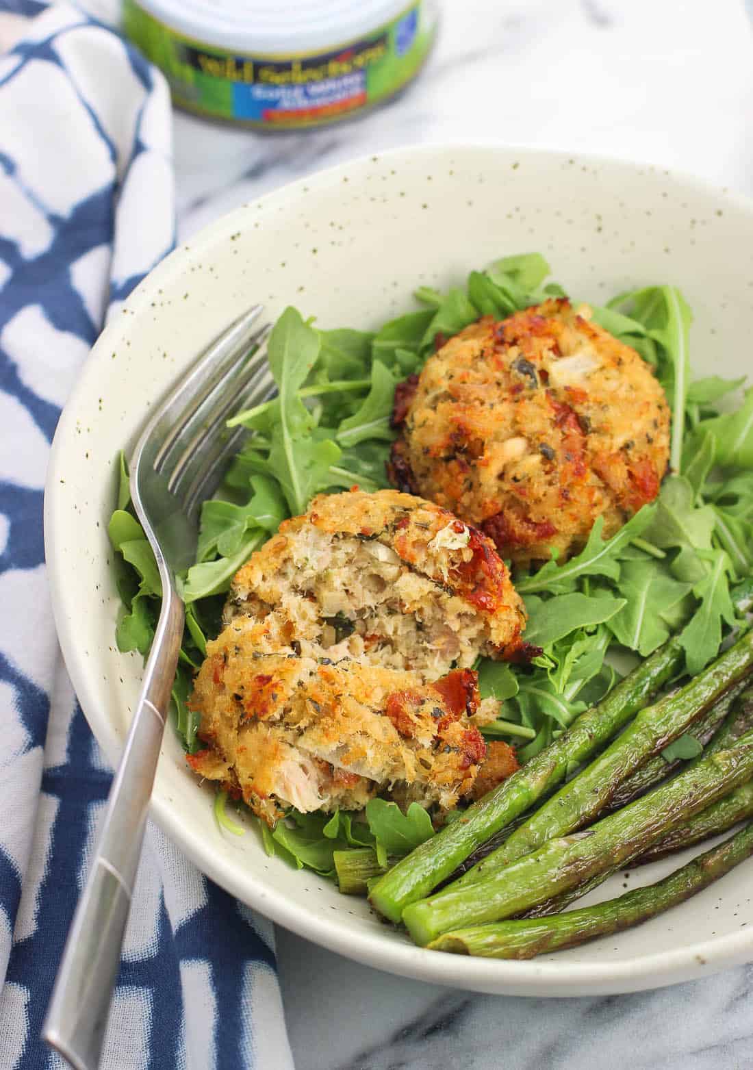 Two tuna cakes on a plate served with arugula and asparagus. One tuna cake is cut in half to show the interior