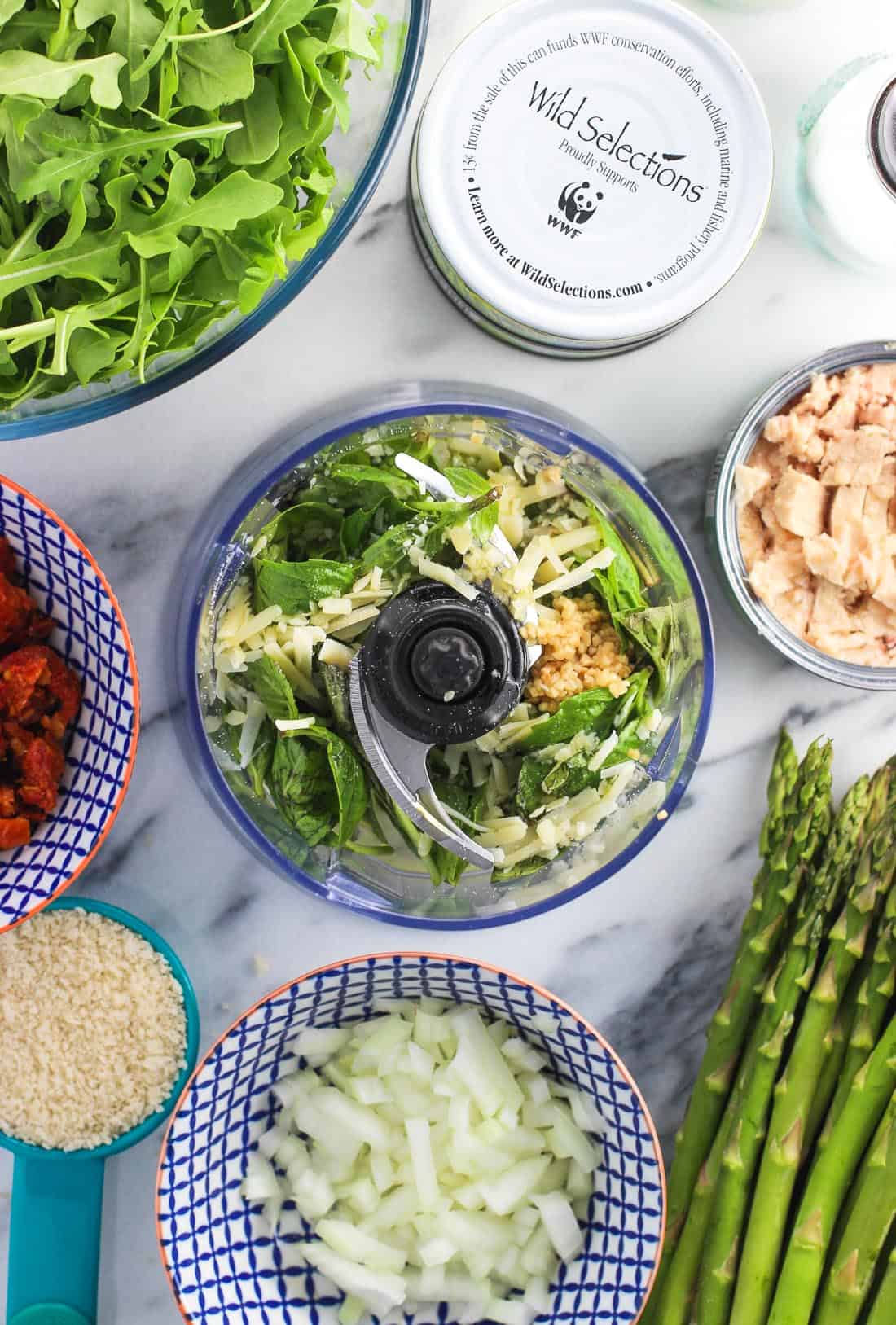 An overhead shot of ingredients in the food processor next to open cans of tuna, a bunch of asparagus, a bowl of arugula, etc.