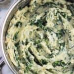 An overhead photo of a large pan filled with mashed potatoes swirled with kale next to glass salt and pepper shakers and a wooden spoon