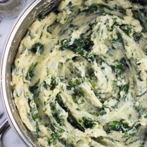 An overhead photo of a large pan filled with mashed potatoes swirled with kale next to glass salt and pepper shakers and a wooden spoon