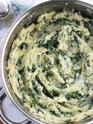 An overhead photo of a large pan filled with mashed potatoes swirled with kale next to glass salt and pepper shakers and a wooden spoon