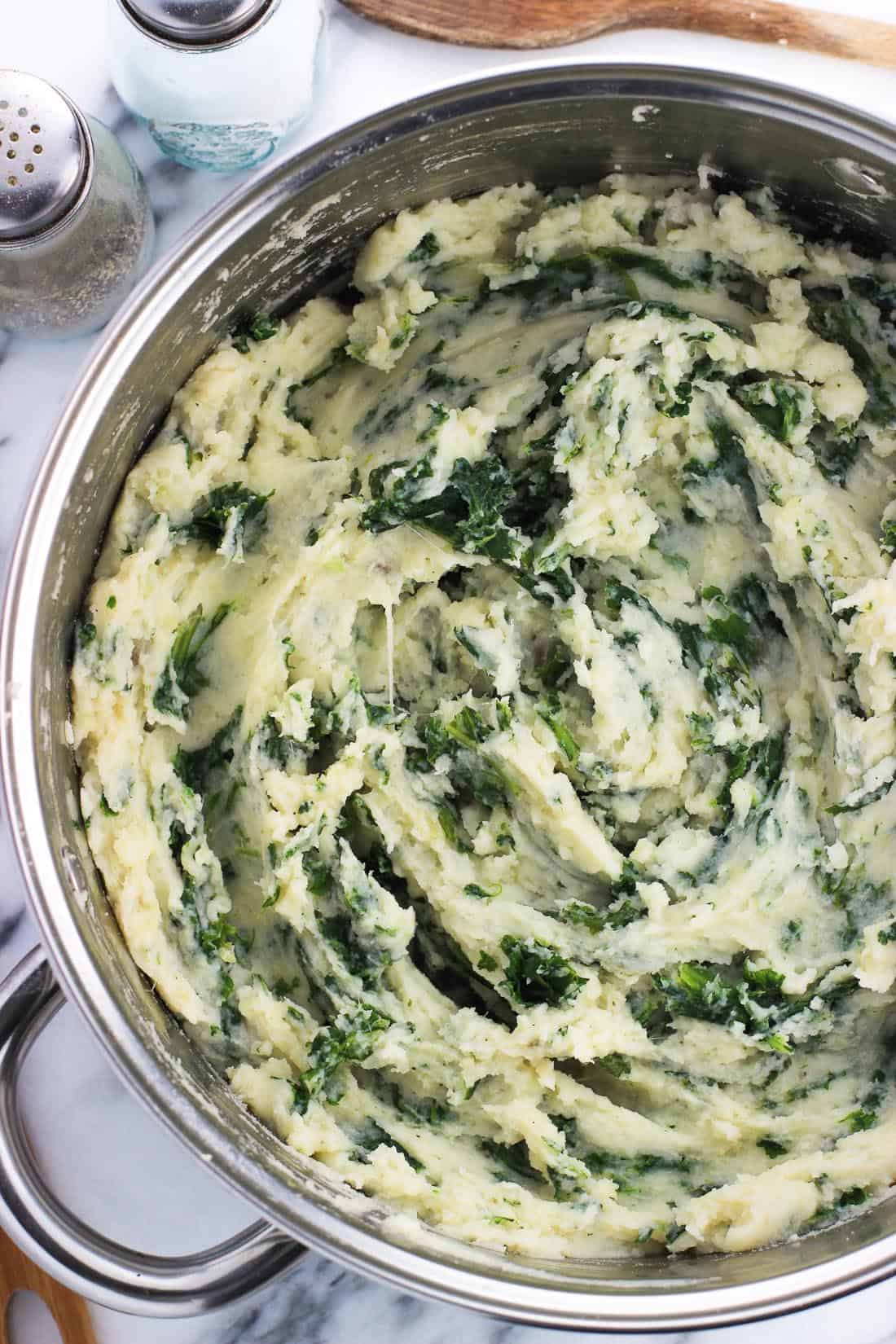 An overhead photo of a large pan filled with mashed potatoes swirled with kale next to glass salt and pepper shakers and a wooden spoon