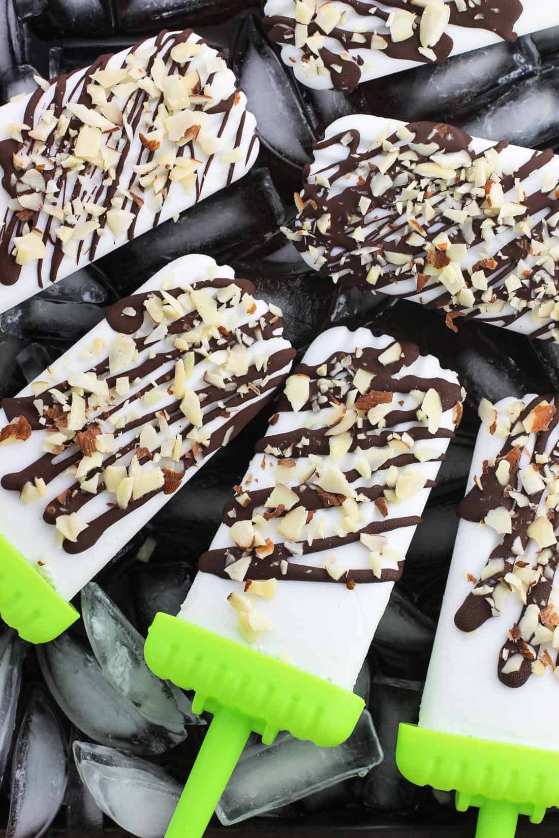 Six coconut popsicles drizzled with chocolate set on an ice cube lined baking sheet