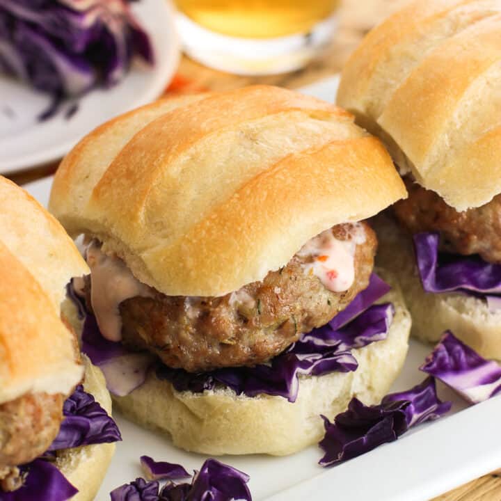 Three pork burger sliders on a rectangular tray, each on slider buns with red cabbage and chili garlic sauce