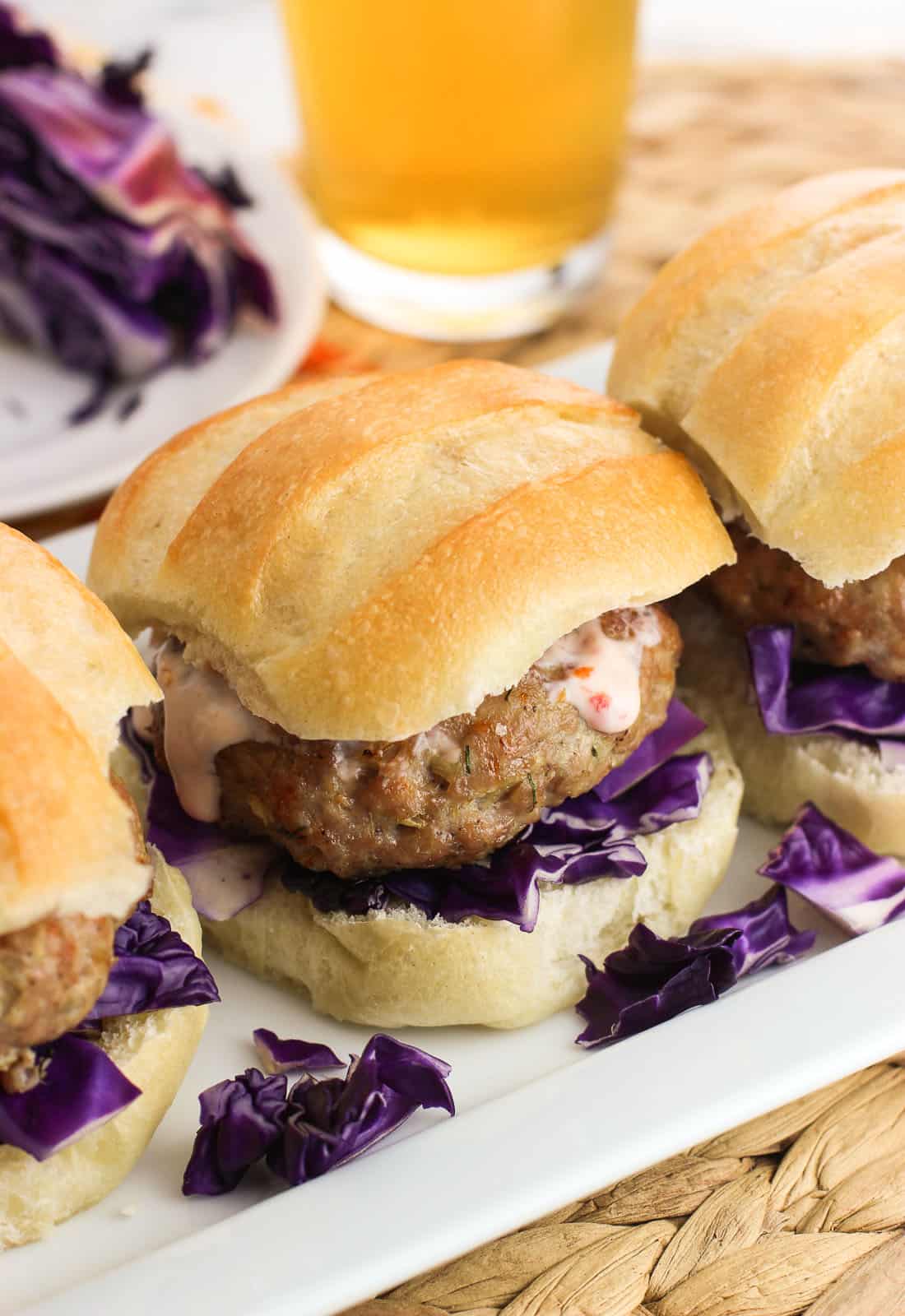 Three pork burger sliders on a rectangular tray, each on slider buns with red cabbage and chili garlic sauce