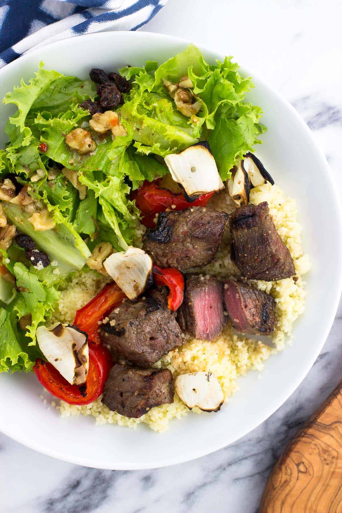 An overhead picture of grilled steak cubes served on couscous with veggies and a side salad, with one of the pieces of steak cut in half