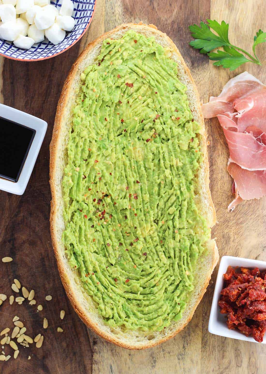 Mashed avocado on a half-loaf of garlic bread on a wooden board surrounded by toppings