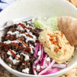 A shallow bowl filled with spiced ground beef, cucumber slices, yogurt sauce, feta, red onions, hummus, and pita