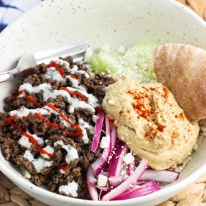 A shallow bowl filled with spiced ground beef, cucumber slices, yogurt sauce, feta, red onions, hummus, and pita