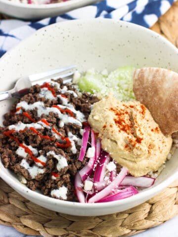 A shallow bowl filled with spiced ground beef, cucumber slices, yogurt sauce, feta, red onions, hummus, and pita