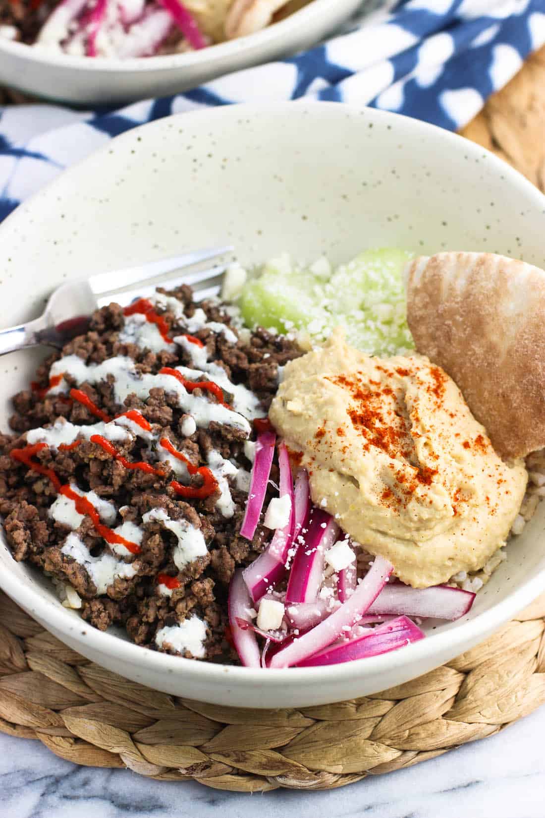 A shallow bowl filled with spiced ground beef, cucumber slices, yogurt sauce, feta, red onions, hummus, and pita