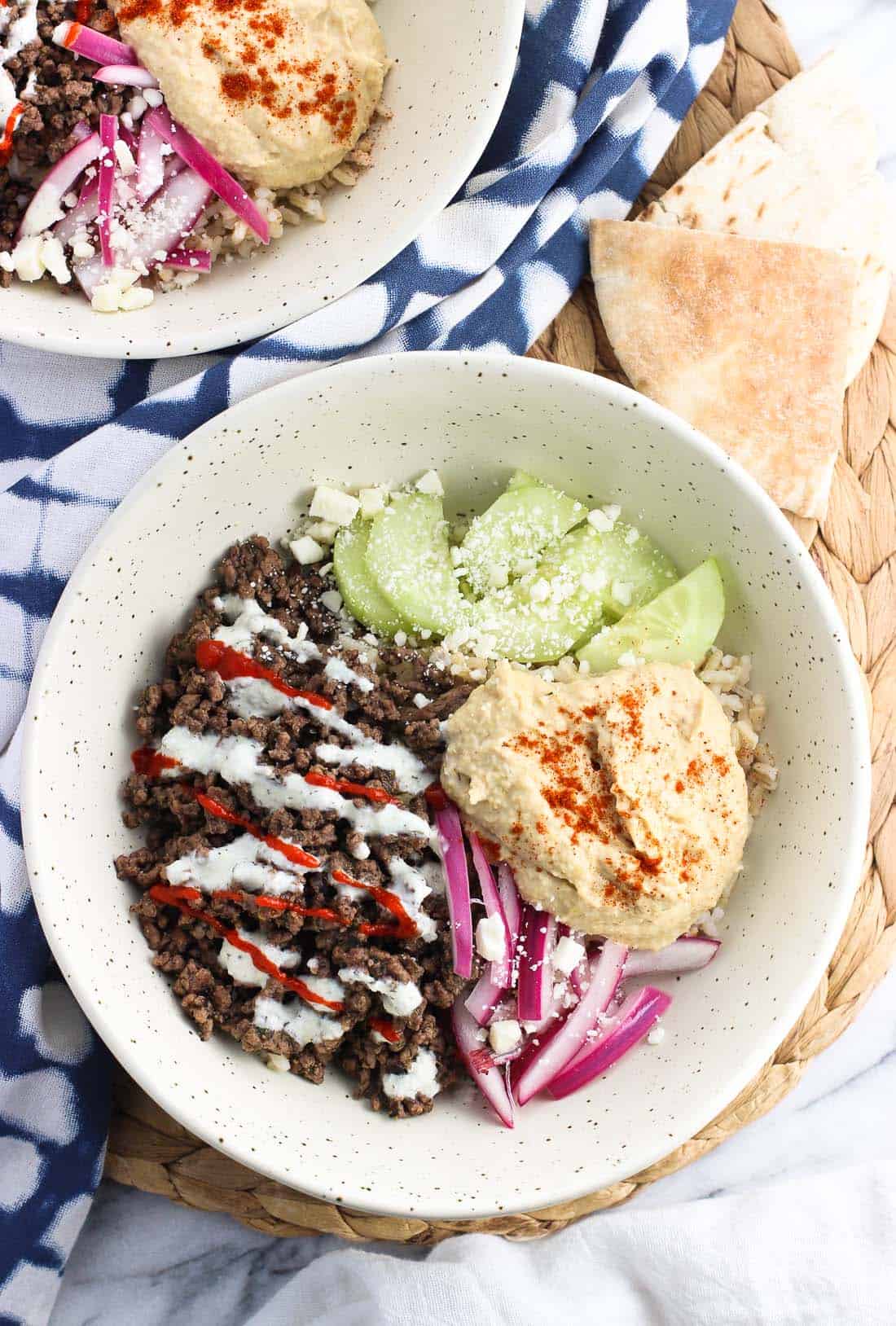 An overhead picture of a rice bowl featuring spiced beef, red onion, hummus, and cucumber slices 