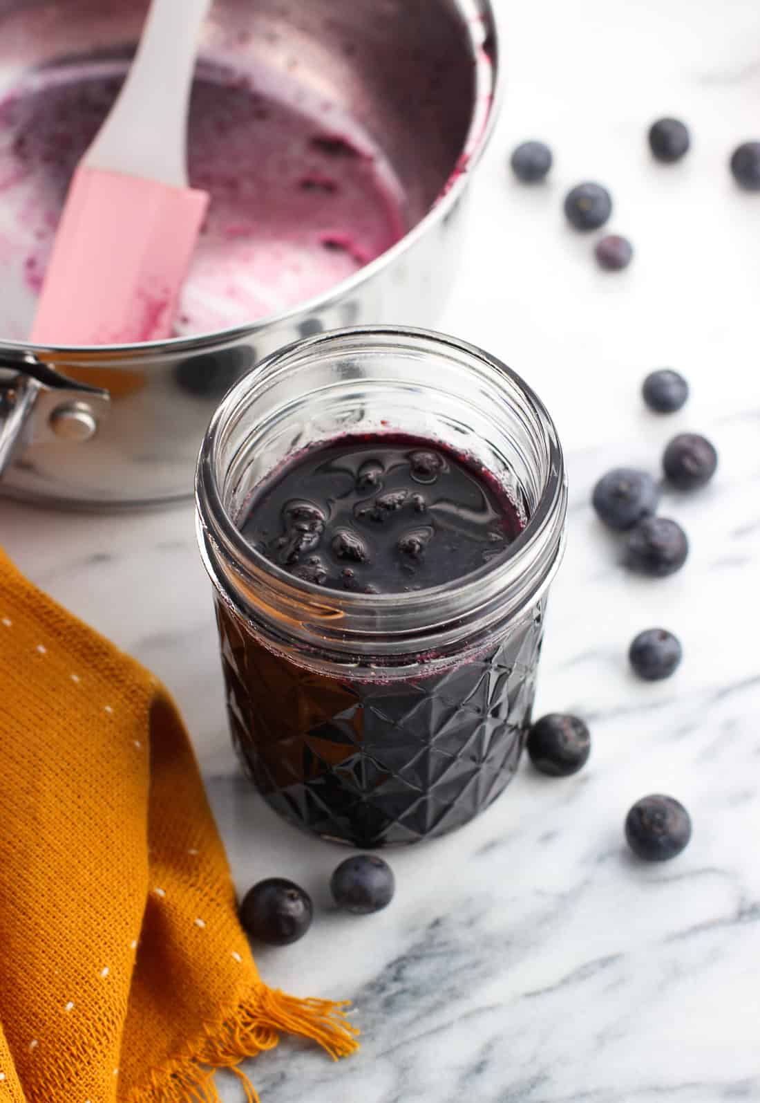 A jar of blueberry syrup next to an empty saucepan and loose blueberries on a marble board