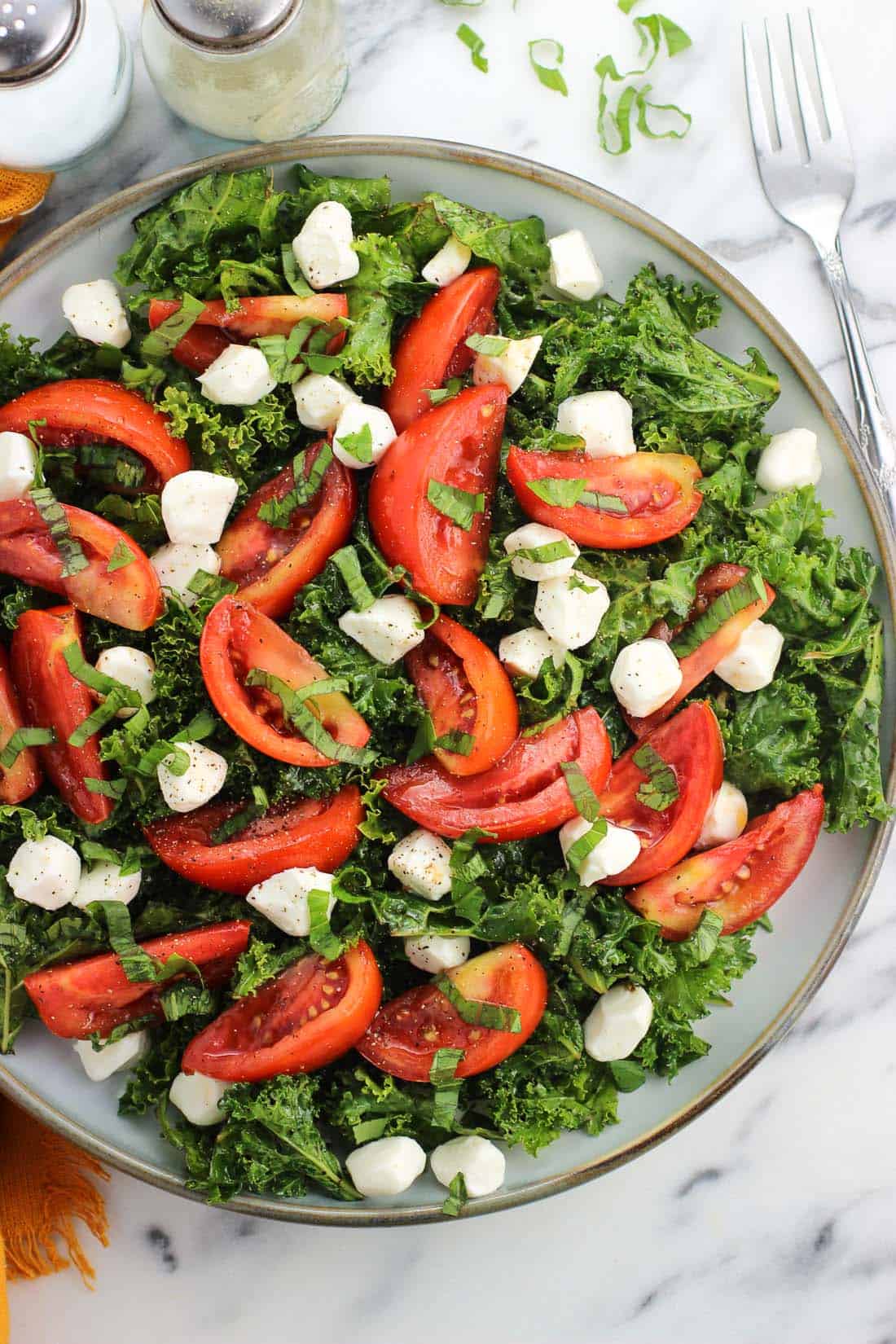 An overhead picture of kale, tomatoes, mozzarella pearls, and basil on a large plate next to a fork and salt and pepper shakers