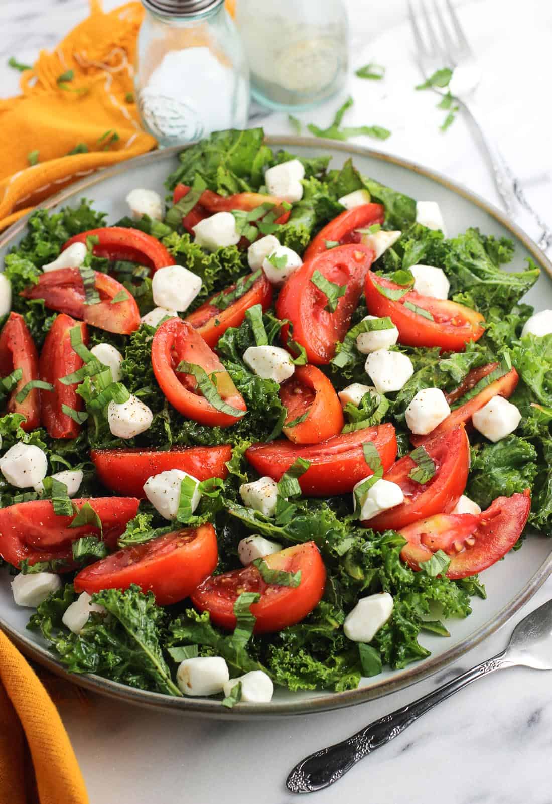 Marinated kale caprese salad on a large plate next to a dish towel, salt and pepper shakers, and forks