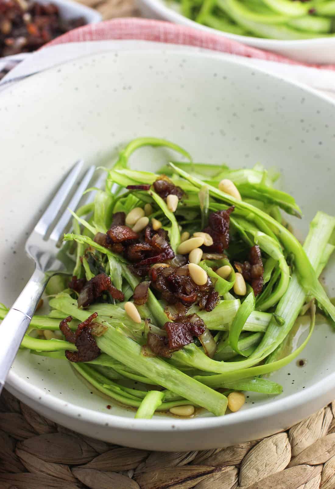 Shaved asparagus in a bowl topped with bacon vinaigrette and pine nuts.