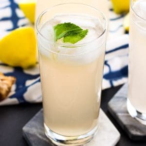 Tall glasses of lemonade on marble coasters next to lemons and ginger root.