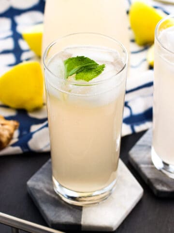 Tall glasses of lemonade on marble coasters next to lemons and ginger root.