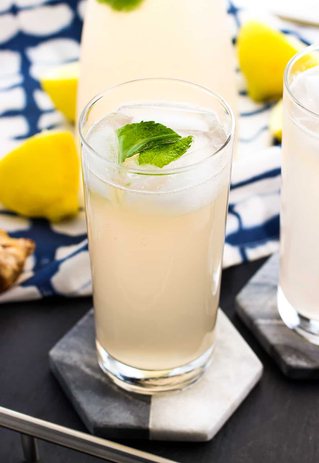 Tall glasses of lemonade on marble coasters next to lemons and ginger root.