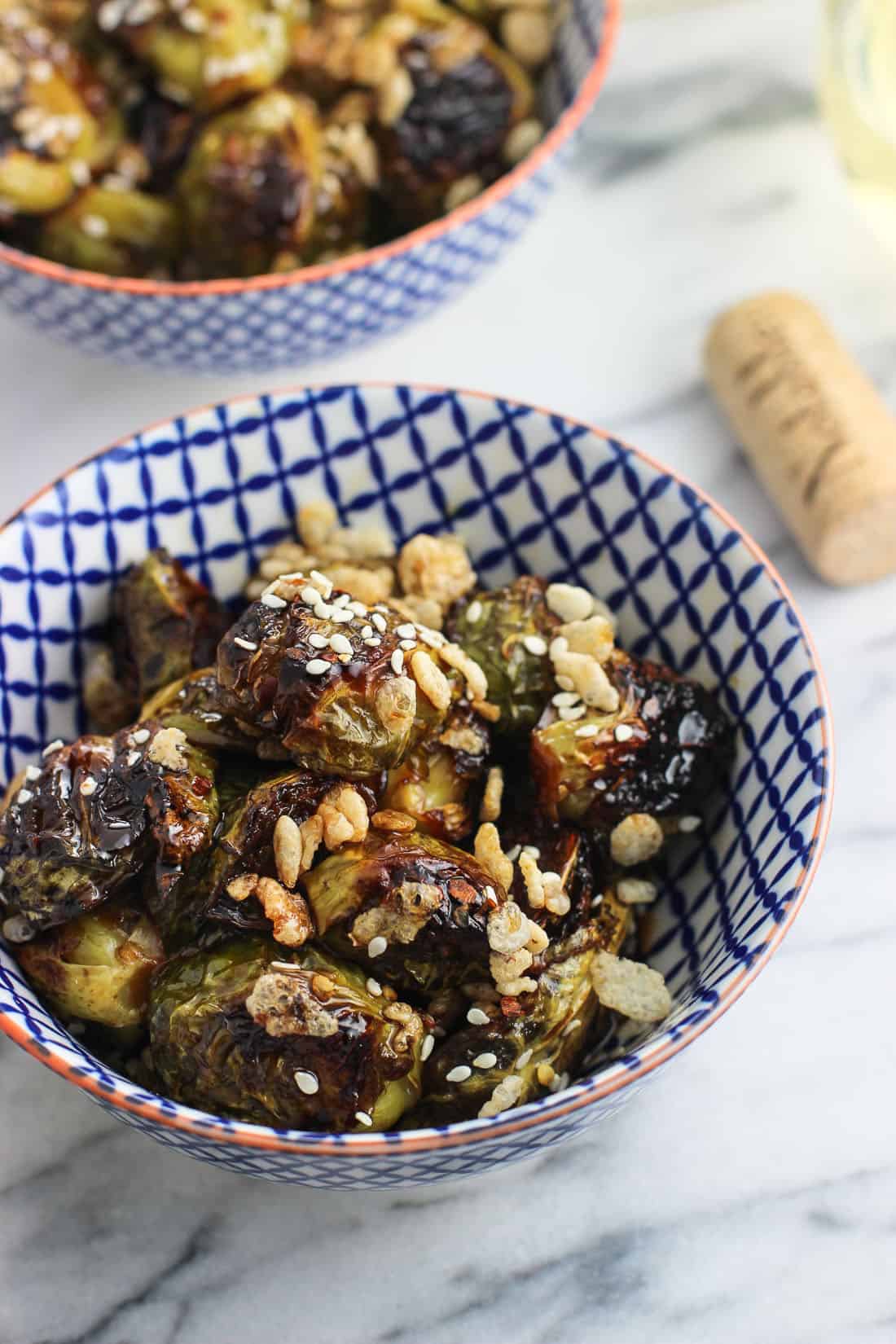 A close-up of a bowl of roasted Brussels sprouts topped with crisped rice with a wine cork and another bowl in the background