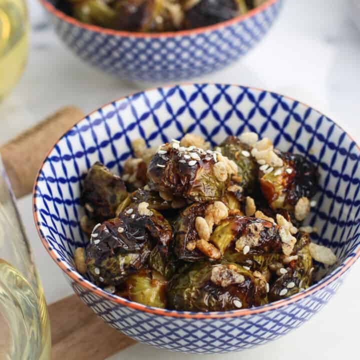 Two bowls of roasted and glazed Brussels Sprouts topped with puffed rice next to two glasses of white wine