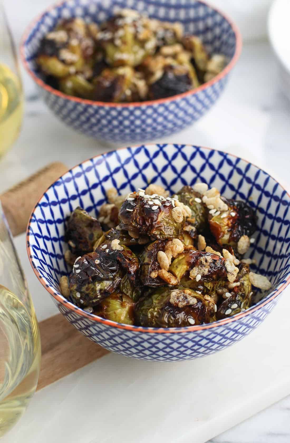 Two bowls of roasted and glazed Brussels Sprouts topped with puffed rice next to two glasses of white wine