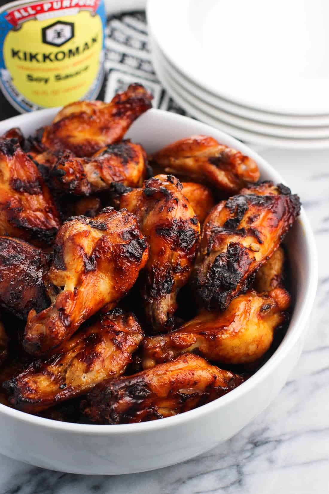 Grilled wings in a ceramic bowl in front of plates and Kikkoman soy sauce bottles.