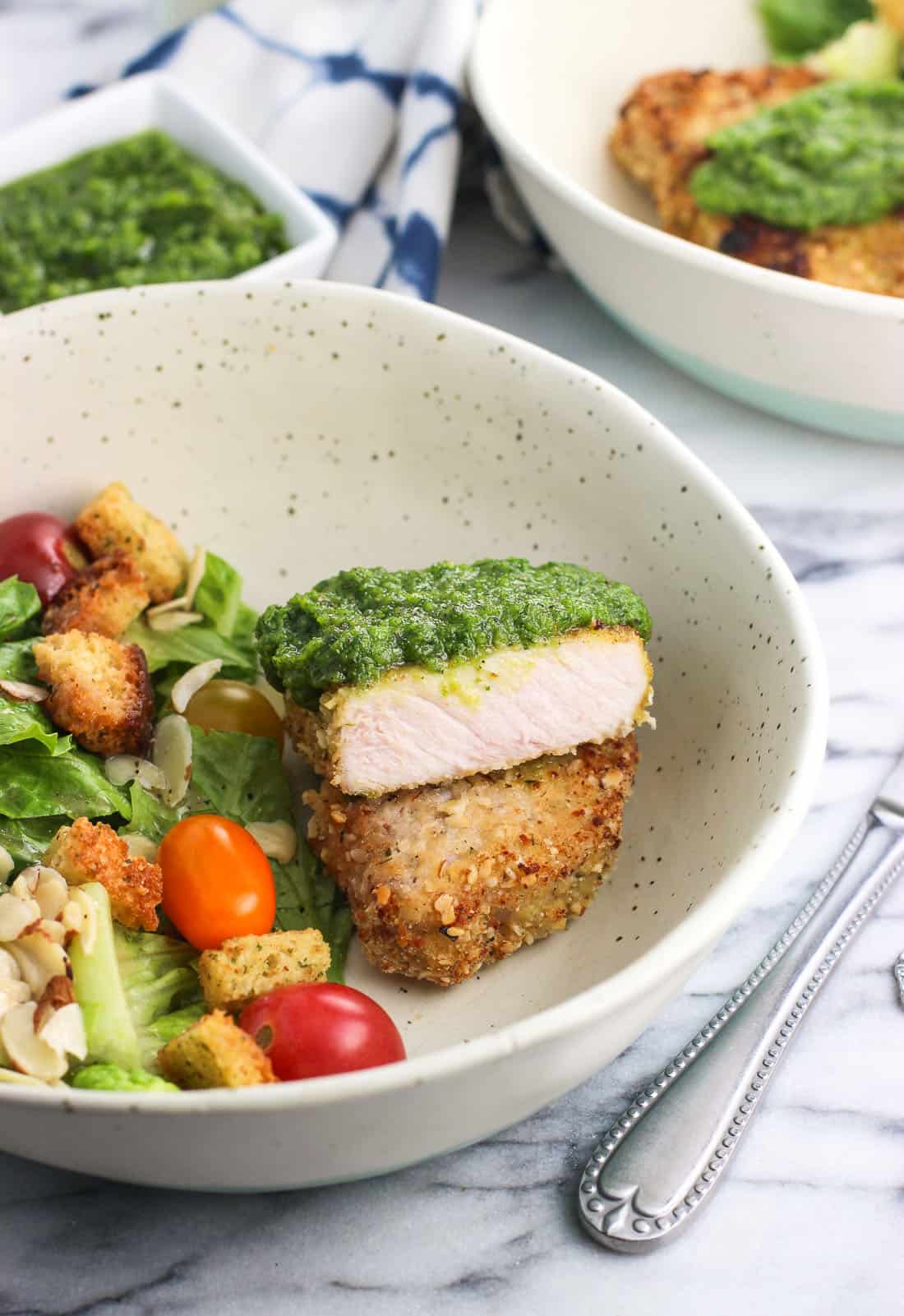 A dish containing a side salad and a pork chop, cut in half and stacked on top of itself, to show the cooked interior topped with pesto