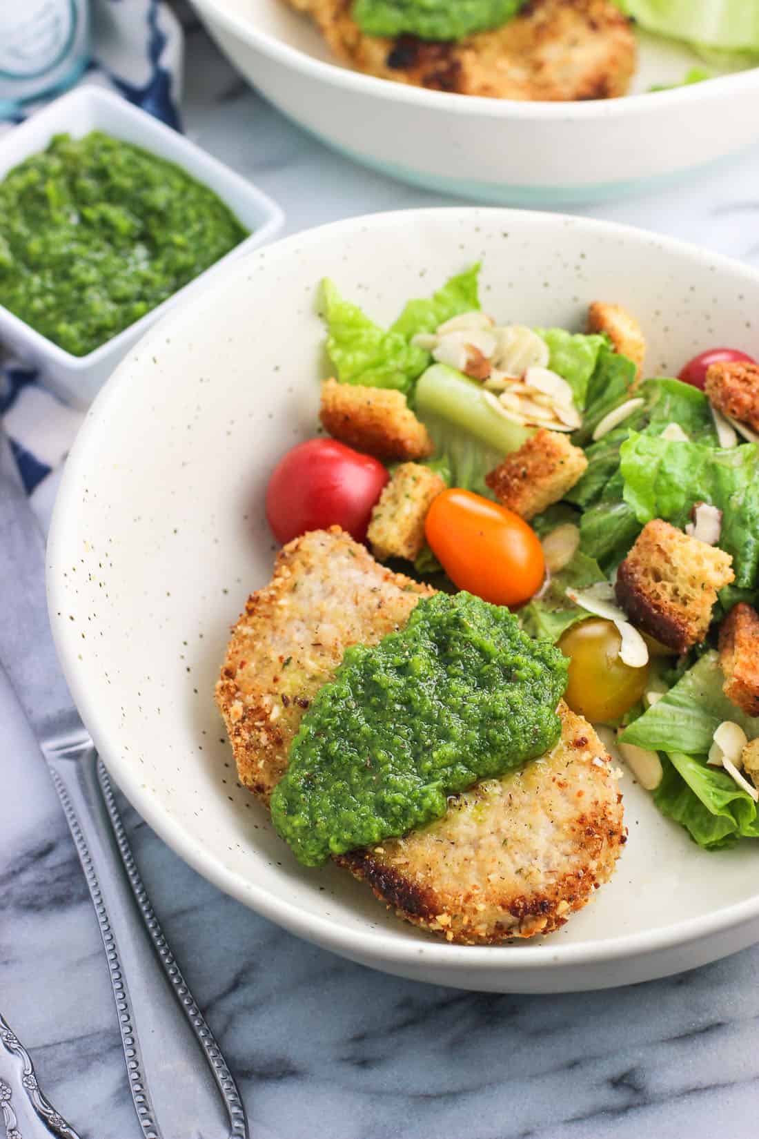 A crusted pork chop on a dish covered in a dollop of parsley pesto and served next to a simple garden salad