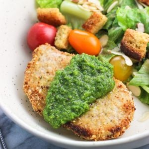 A crusted baked pork chop on a dish served with a dollop of pesto next to a side salad