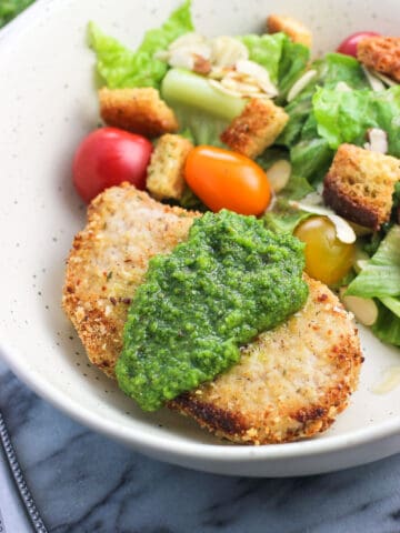 A crusted baked pork chop on a dish served with a dollop of pesto next to a side salad