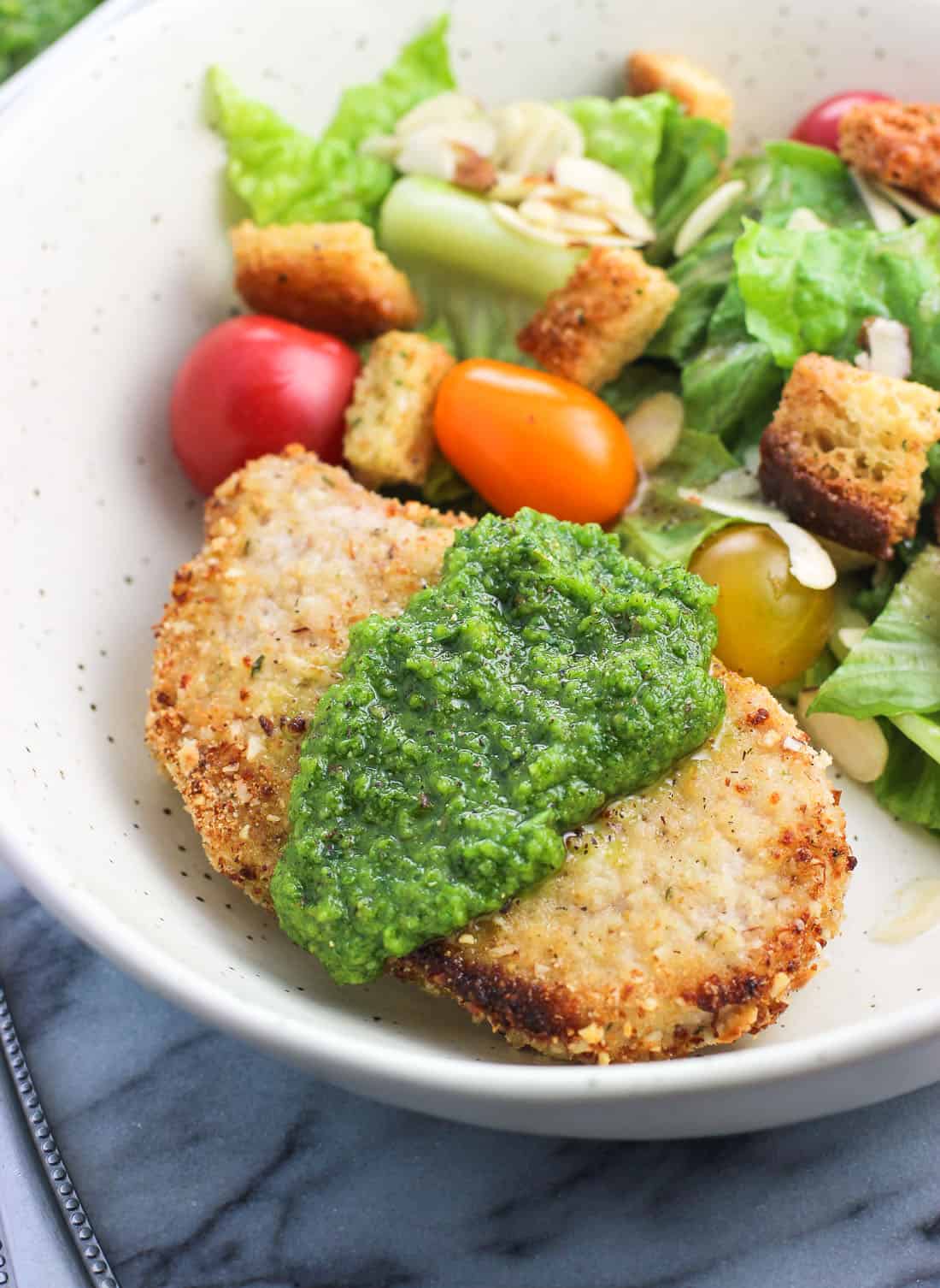 A close-up of a pork chop serving topped with pesto next to a garden salad