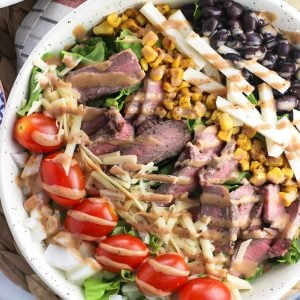 A steak salad in a large bowl with many toppings and drizzled with BBQ ranch dressing.