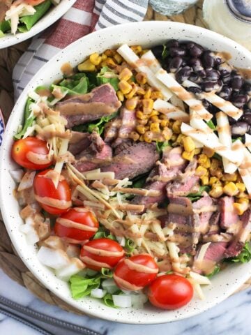 A steak salad in a large bowl with many toppings and drizzled with BBQ ranch dressing.