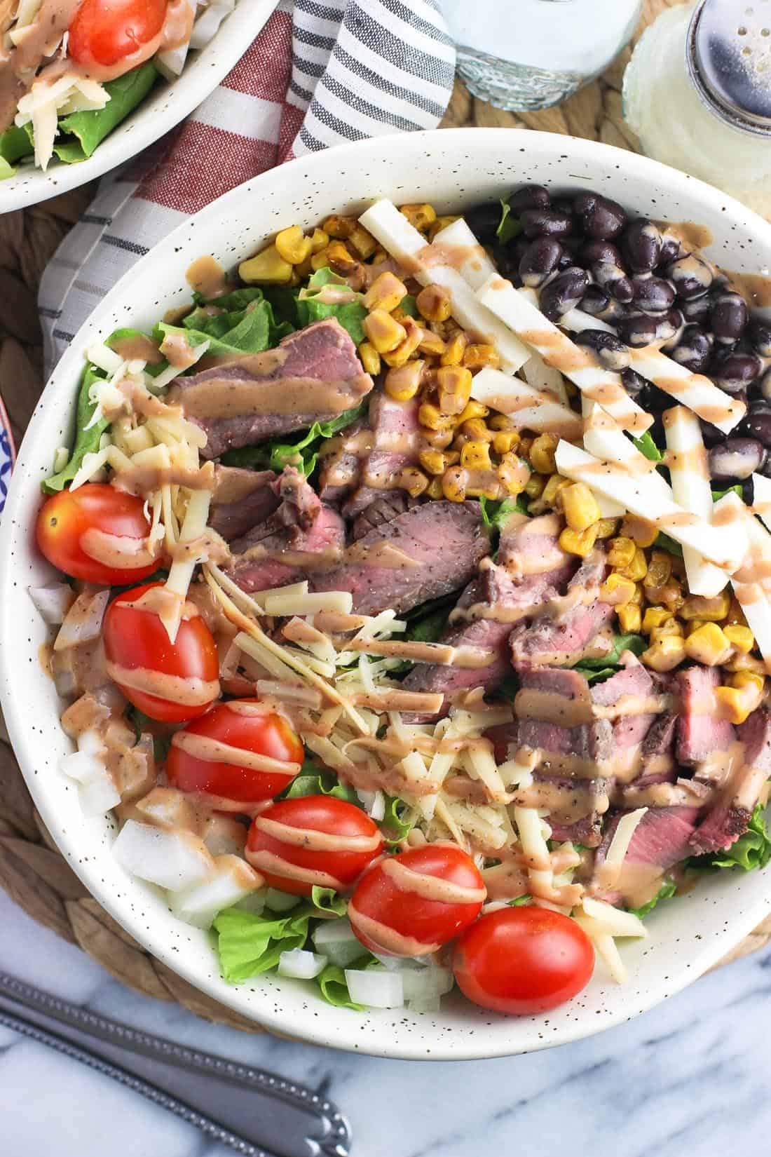 A steak salad in a large bowl with many toppings and drizzled with BBQ ranch dressing.